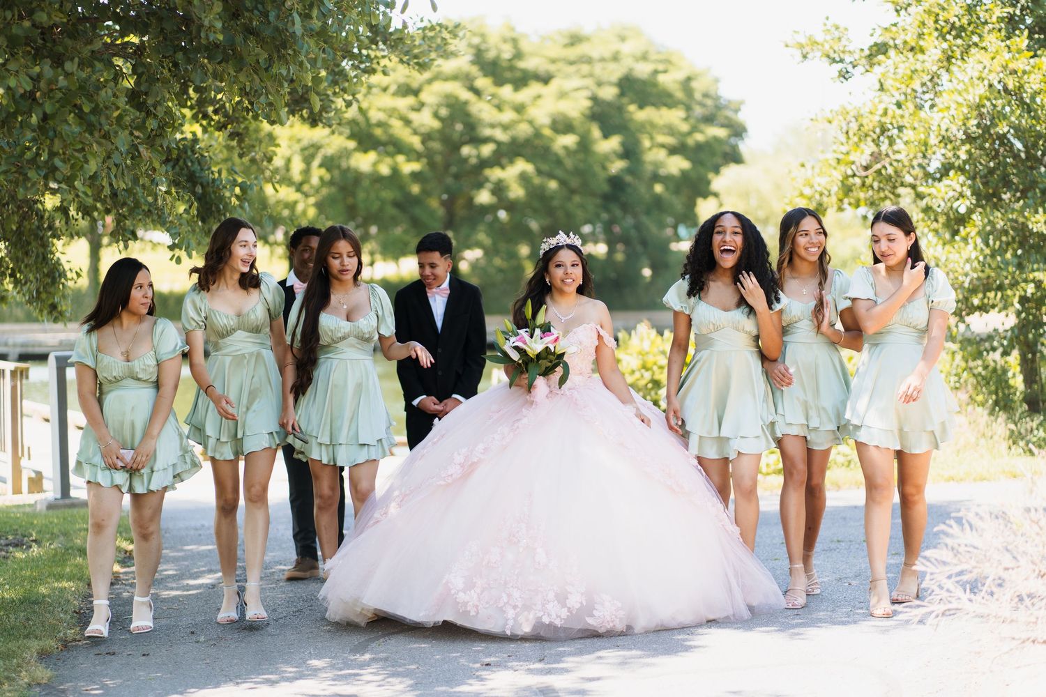 A bridal party in mint green dresses walks together on a sunny path surrounded by trees.