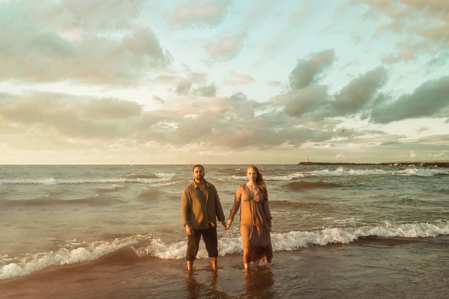 Romantic couple standing in shallow ocean waves during golden hour sunset.