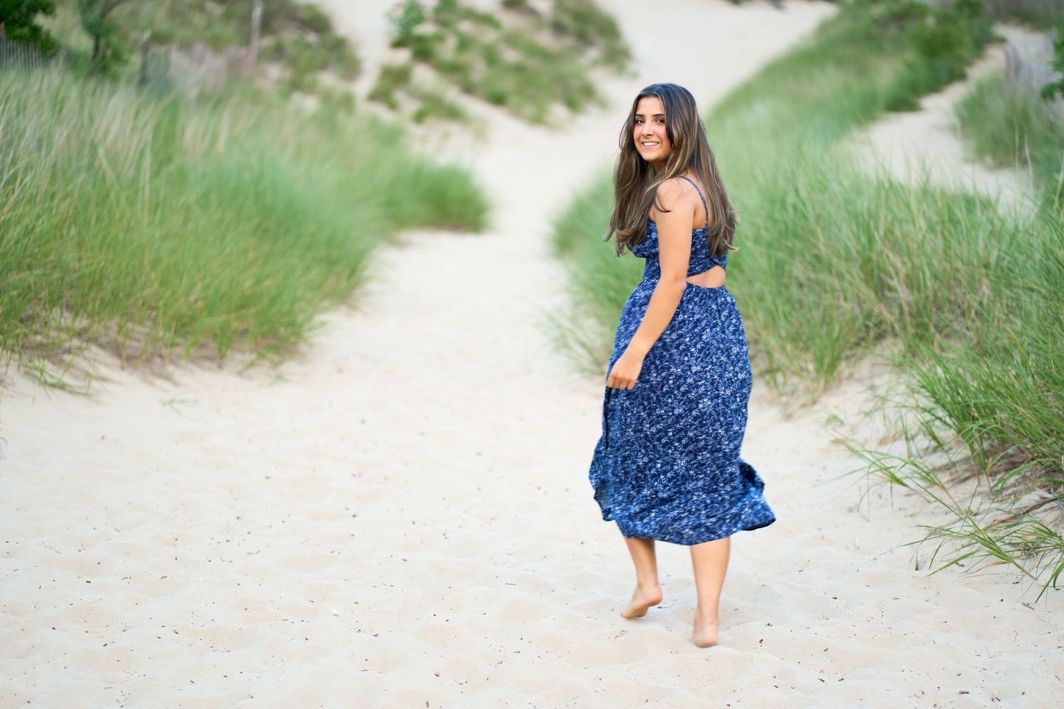 Woman in blue polka dot dress walks barefoot along sandy beach path on sunny summer day.
