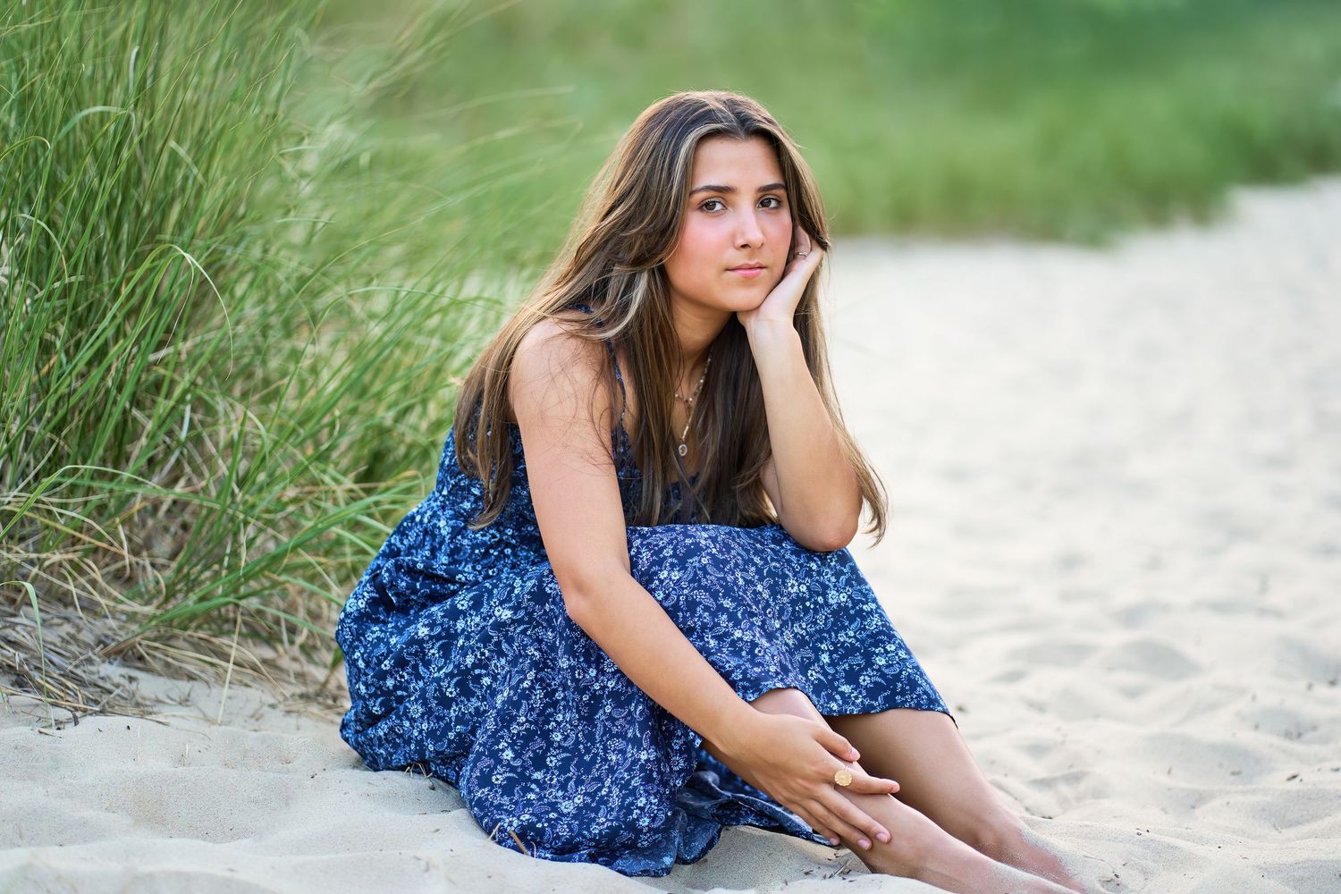 A woman in a blue floral dress sits thoughtfully on a sandy beach path surrounded by beach grass.