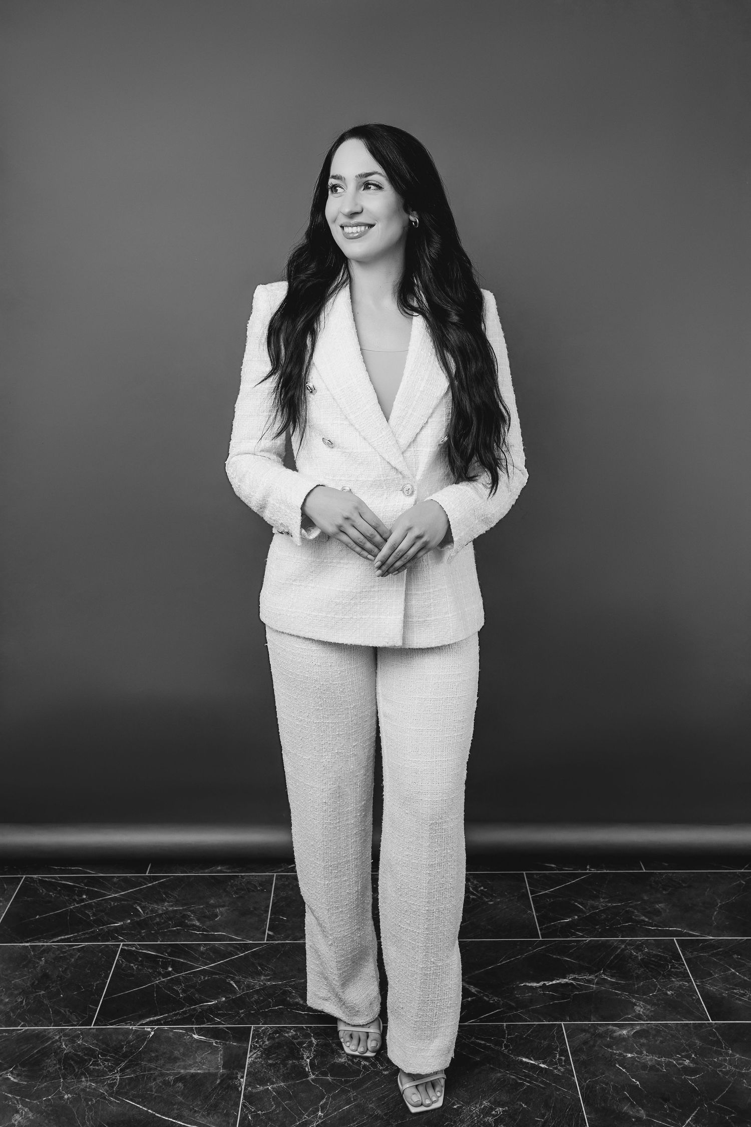 A professional wearing a light-colored pantsuit stands confidently against a dark background in a black and white photo.