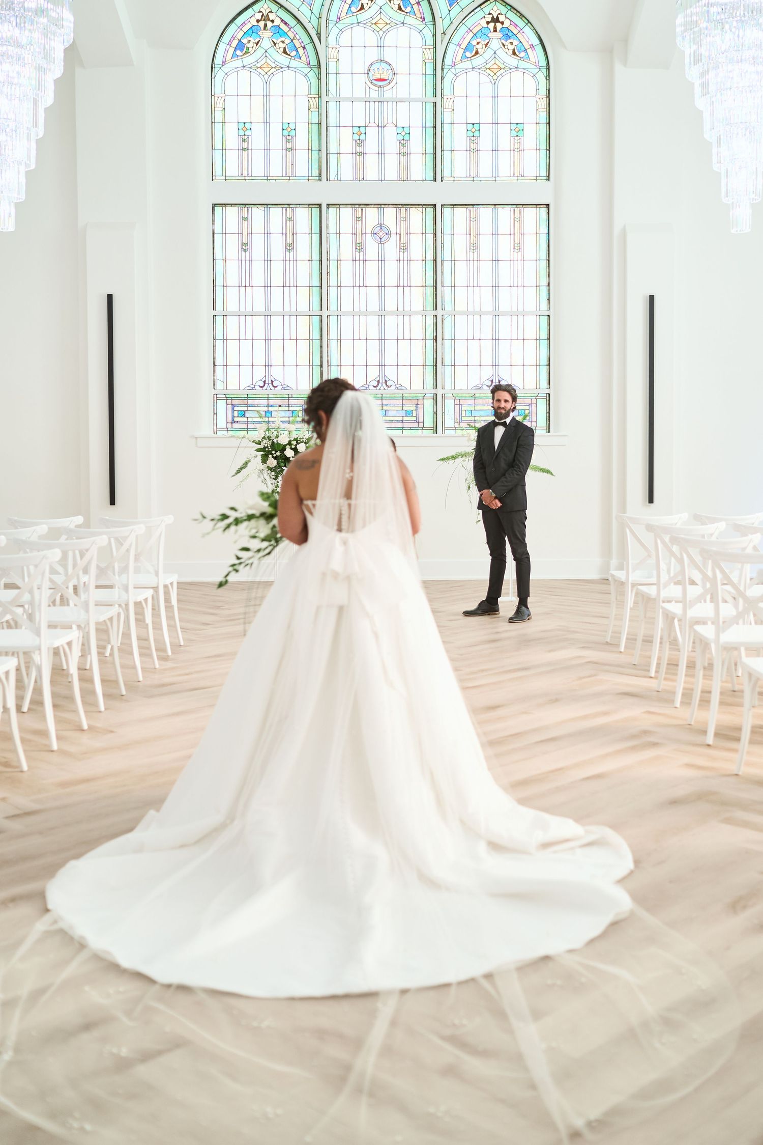 A stunning bride in a flowing white gown stands before beautiful stained glass windows in an elegant chapel.