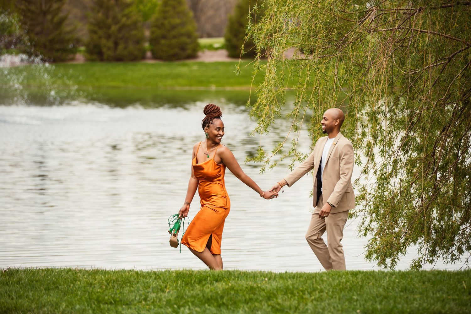 Couple in orange dress and beige suit holding hands while walking by a lake under willow trees.