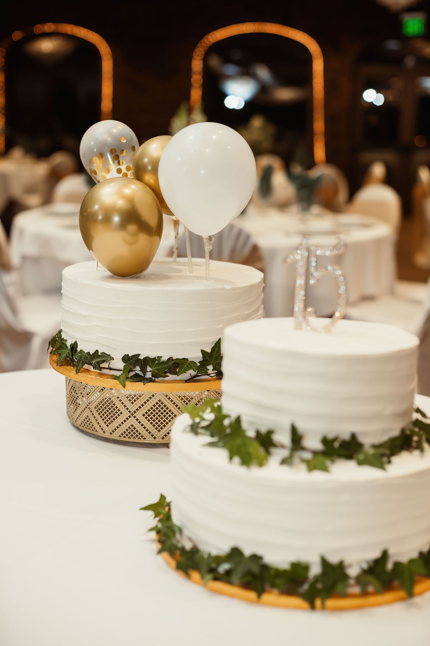 White tiered wedding cakes decorated with gold balloons and greenery garlands on display.