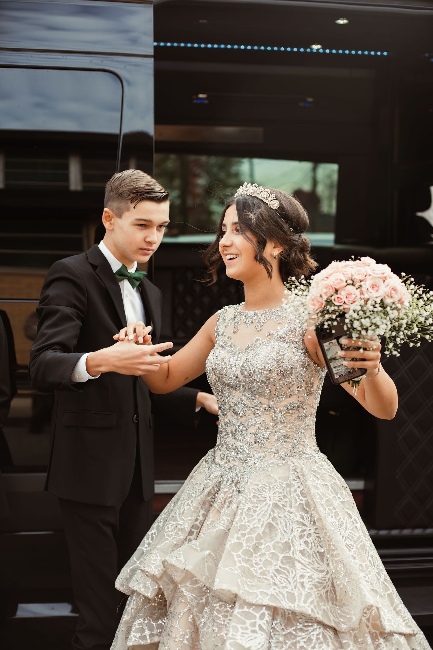 A joyful wedding moment captured outside a black vehicle with a bouquet of pink flowers.