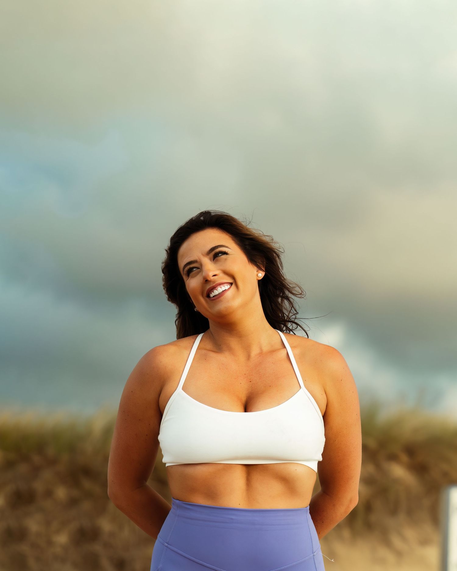 Woman in workout clothing smiles joyfully while posing outdoors on a cloudy day.