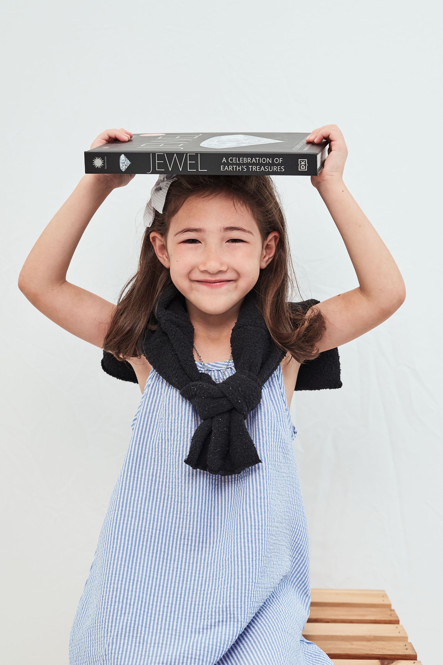 A child in a striped blue dress and black scarf holds a book above their head while smiling.