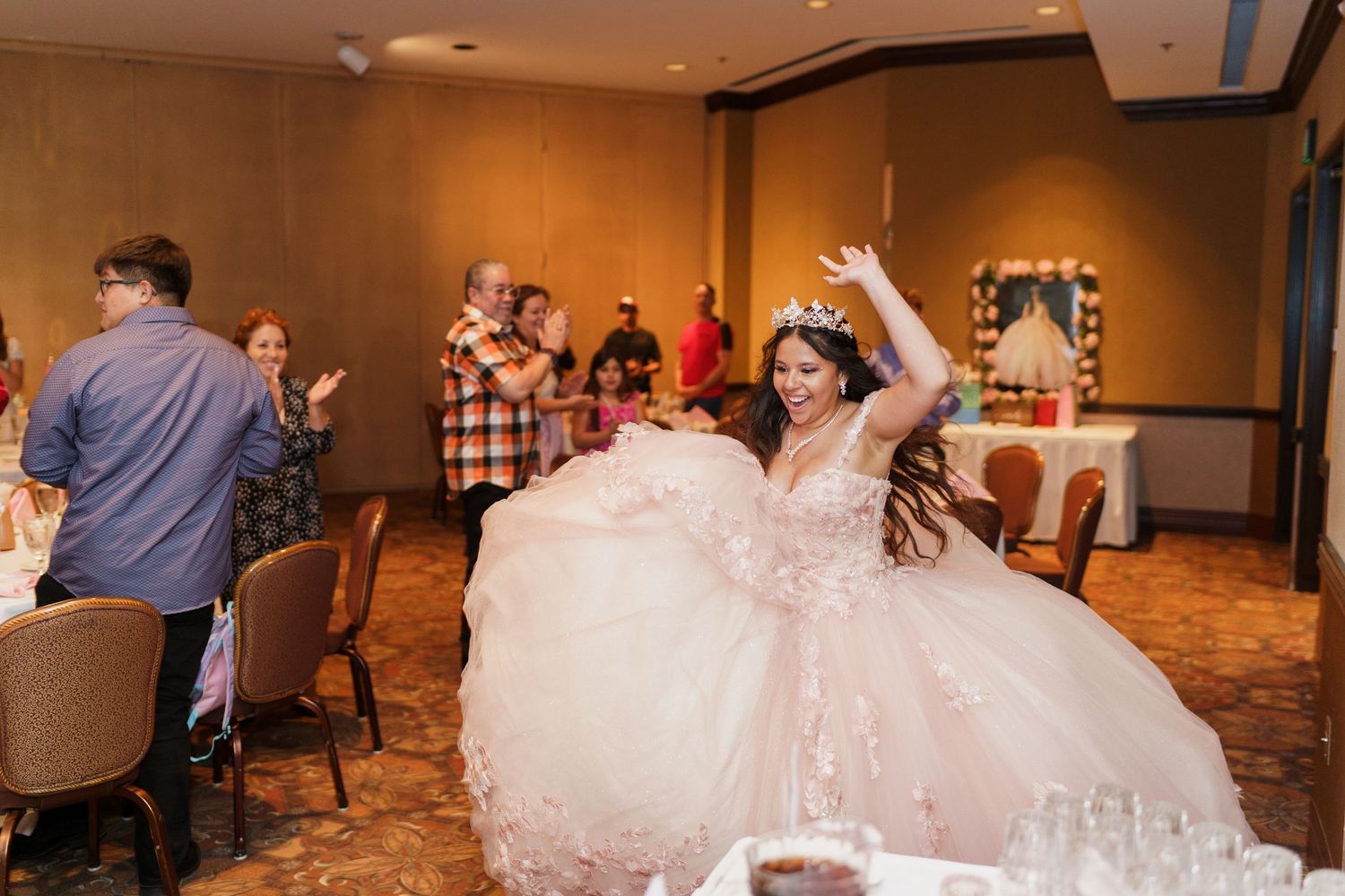 A person in a flowing blush pink quinceanera gown raises their arm joyfully during a celebration in an event hall.