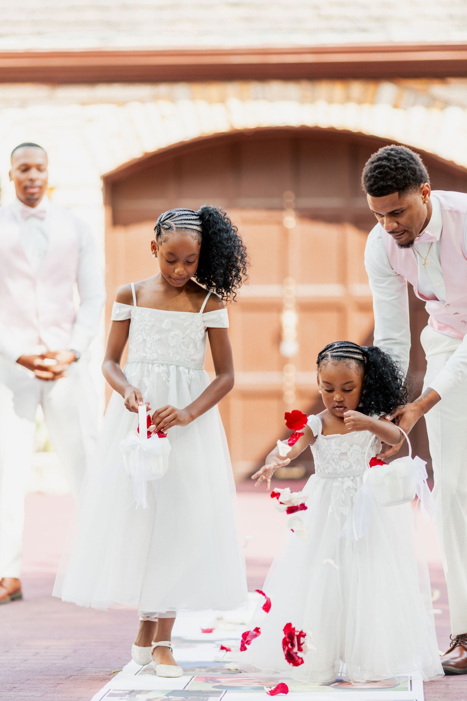 Young children in white dresses scatter rose petals down an aisle during a wedding ceremony.