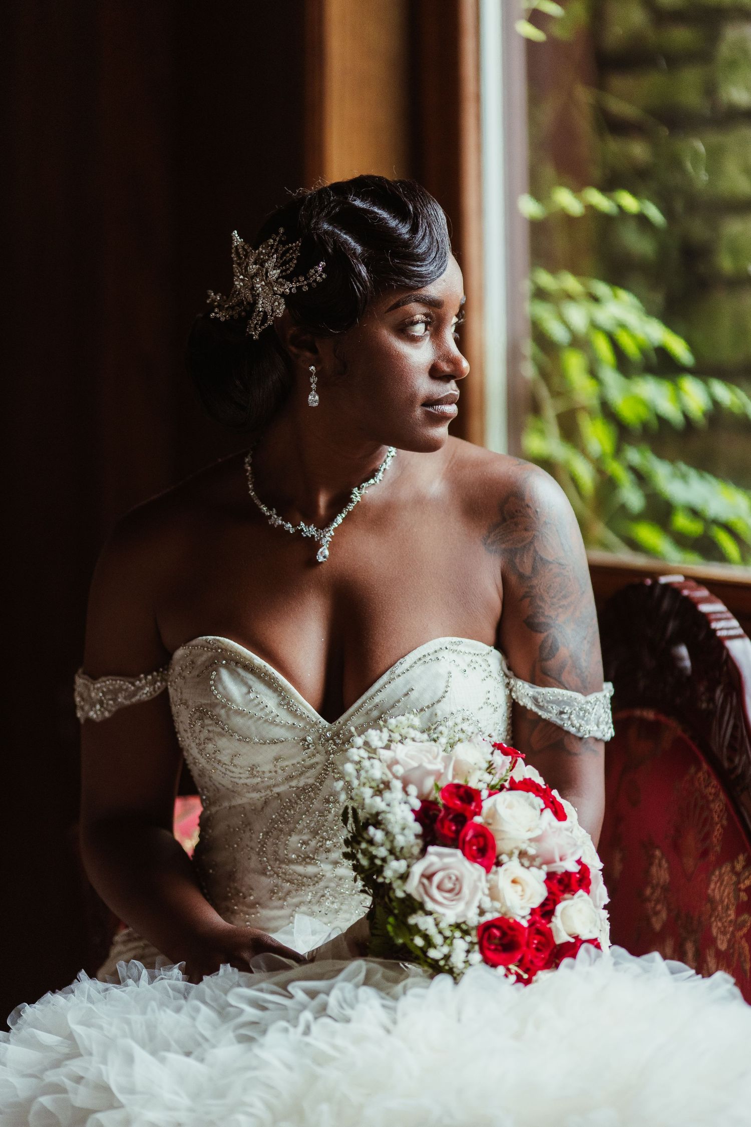 Bride in embellished strapless gown sits by window holding red and white floral bouquet.