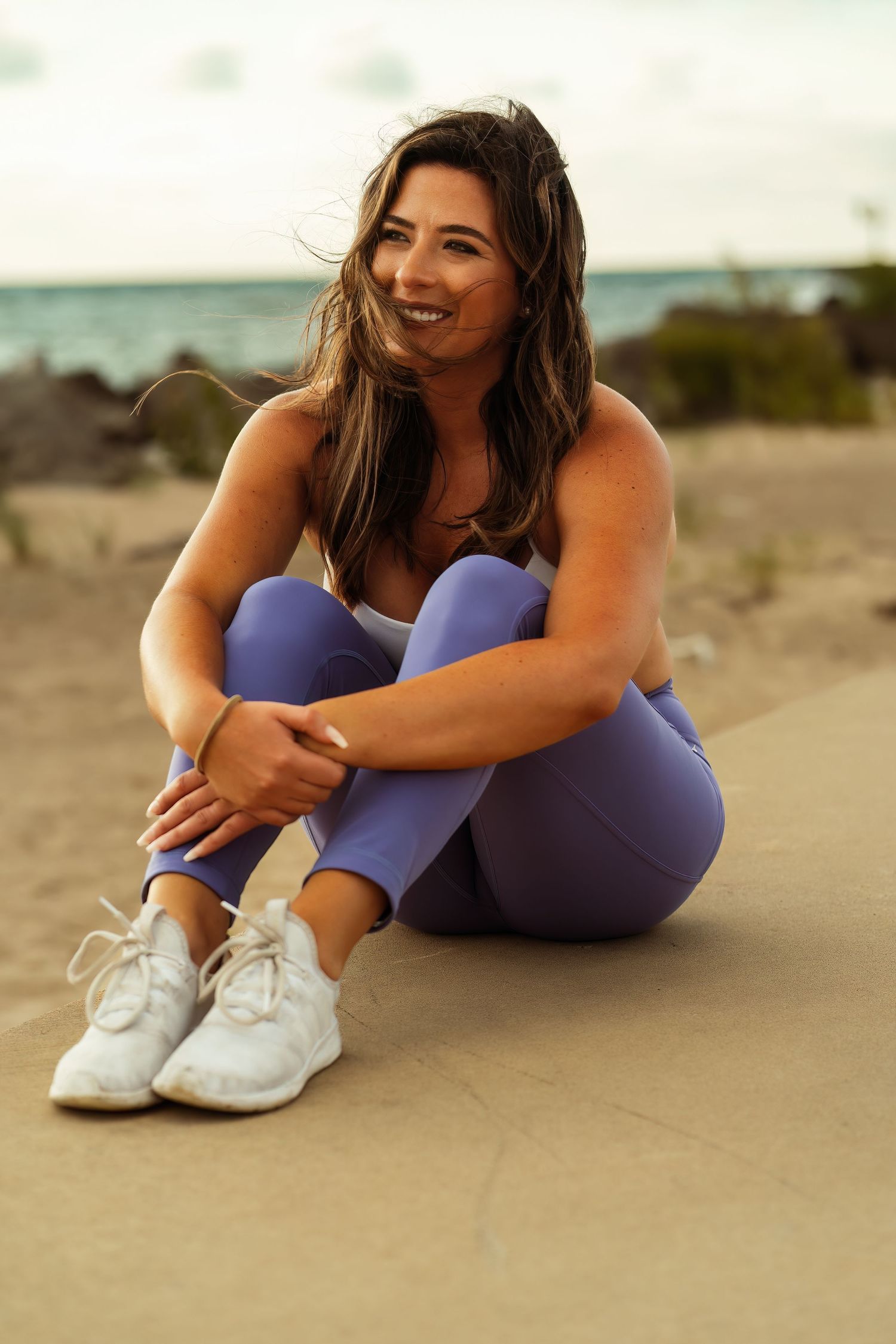 Athletic woman in workout clothes sits on beach with ocean and rocks in background.