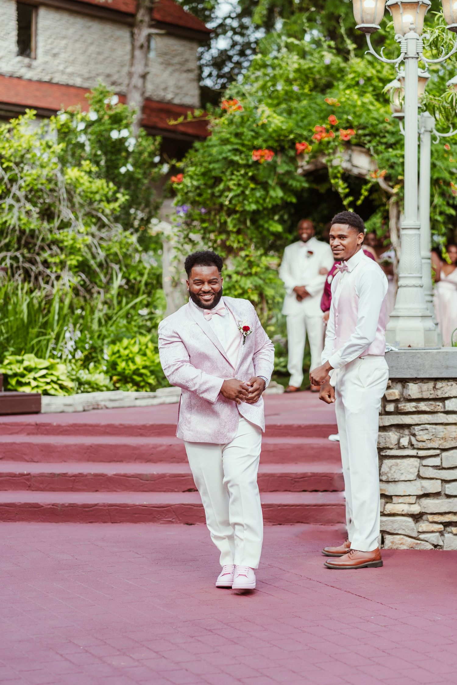 Groomsmen in light pink and white suits walk down red-carpeted outdoor steps.