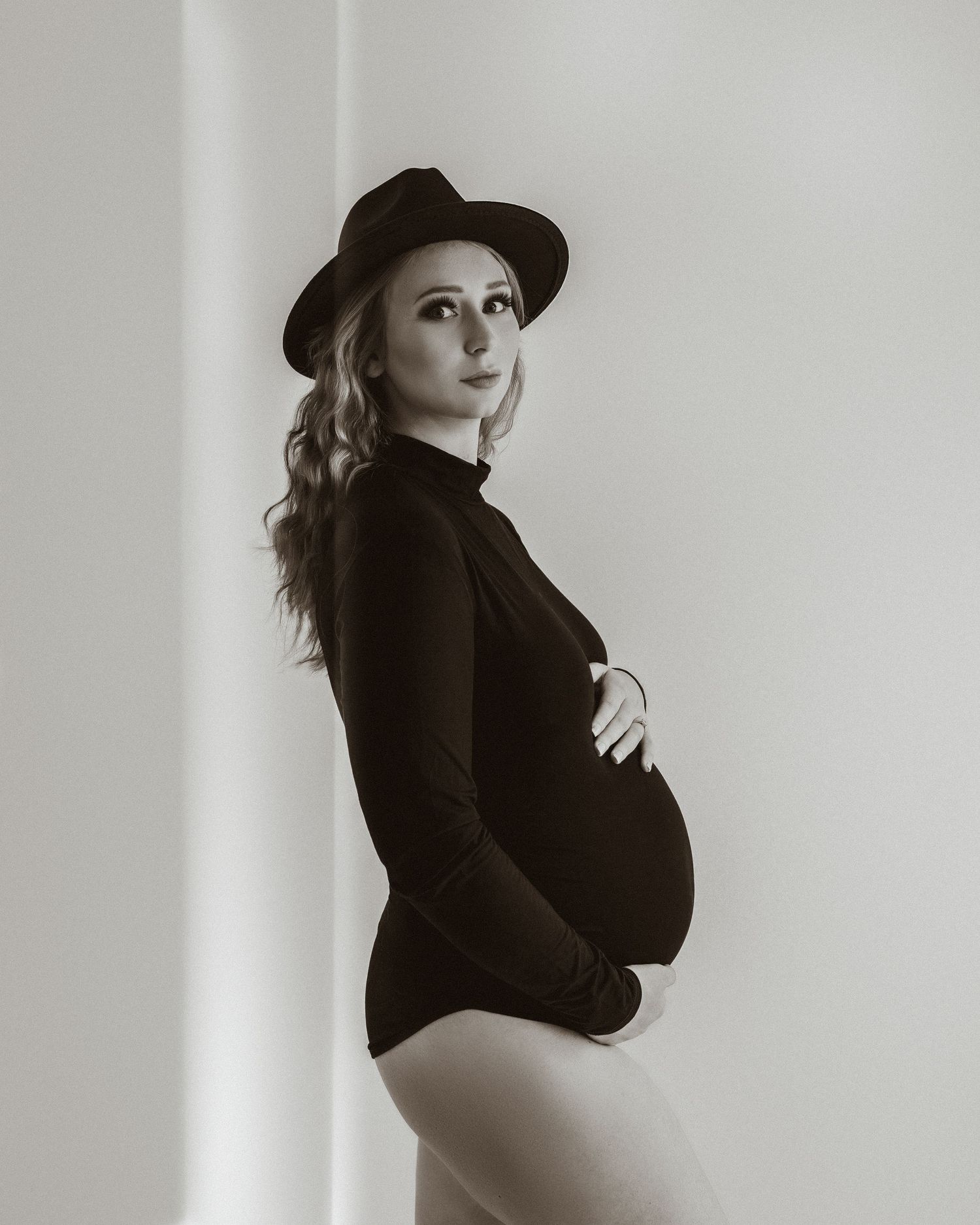 Black and white maternity portrait with dramatic hat and classic styling.
