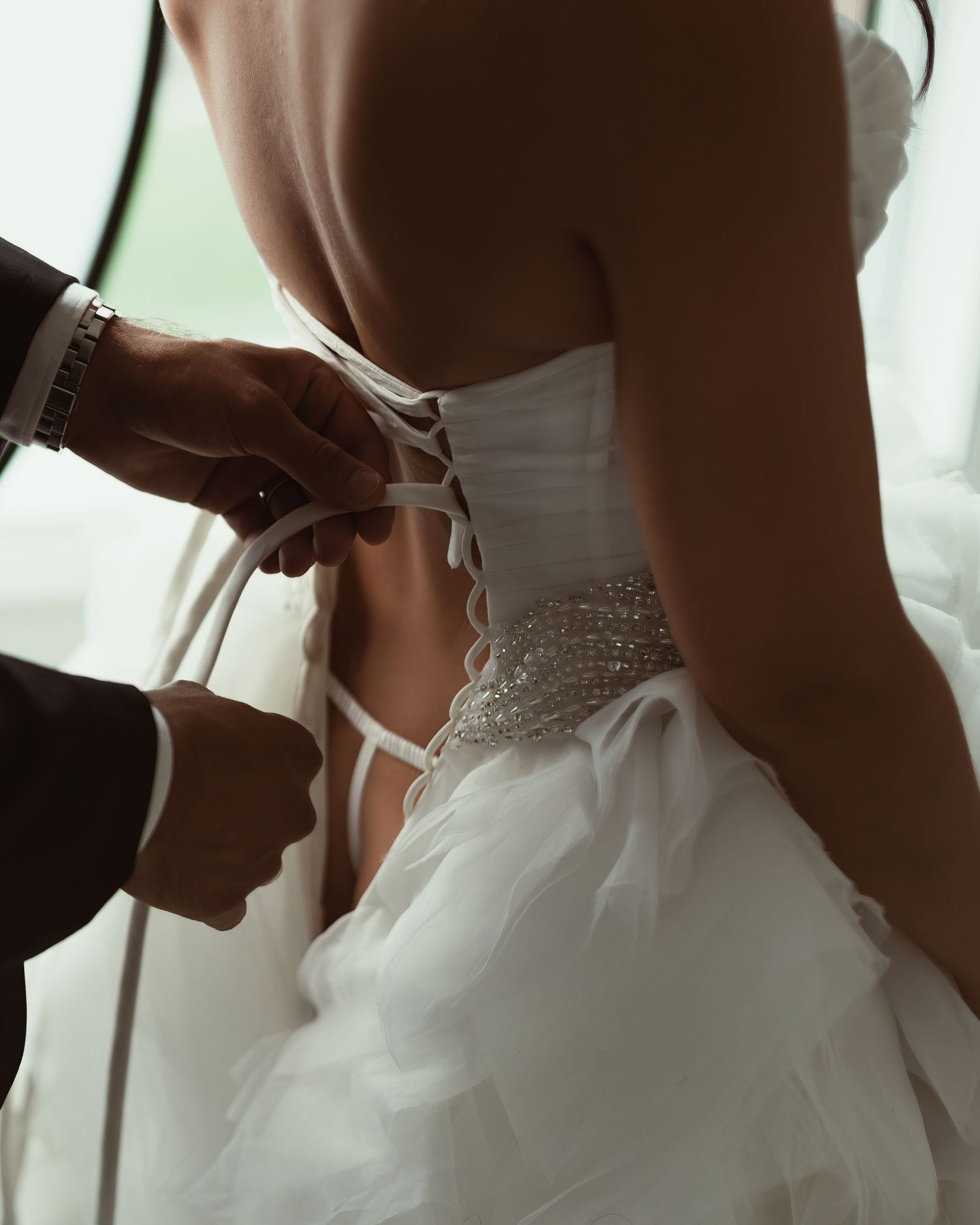Person helping to lace up the back of an ornate white wedding dress with ruched detailing.