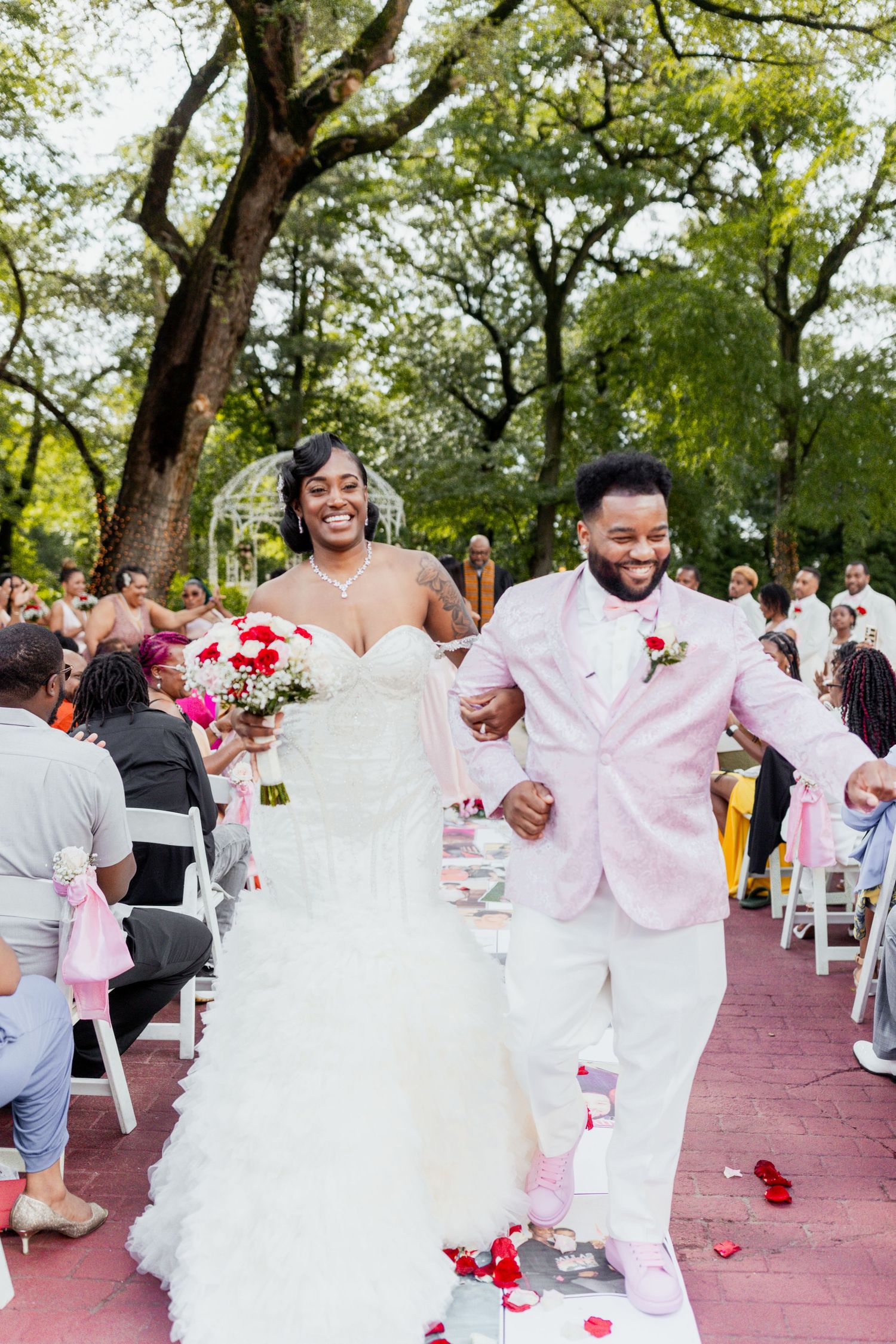 Newlyweds joyfully walk down the red aisle after their outdoor wedding ceremony while guests celebrate.
