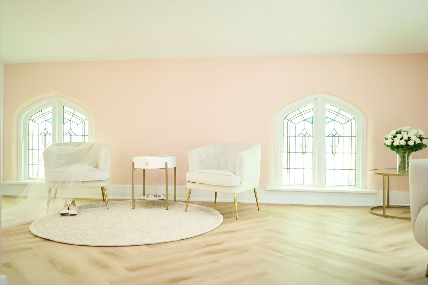 Bright wedding venue interior with white chairs, round rug, and arched windows in soft pink pastel lighting.
