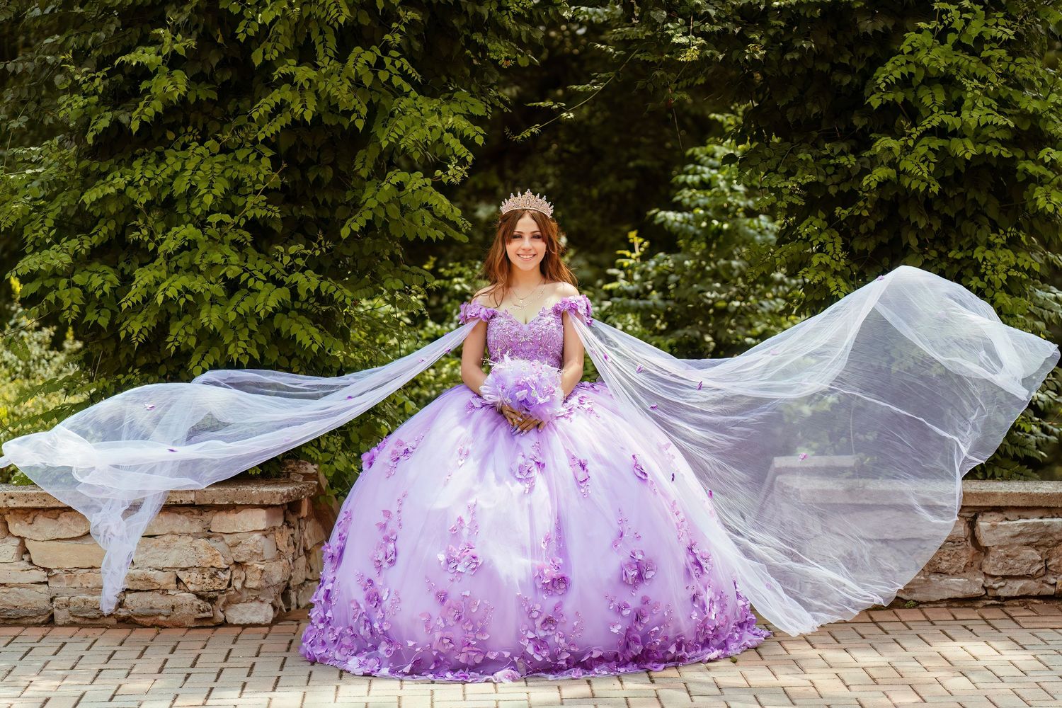 A flowing purple floral gown with a long white veil creates an ethereal scene against a garden stone wall backdrop.