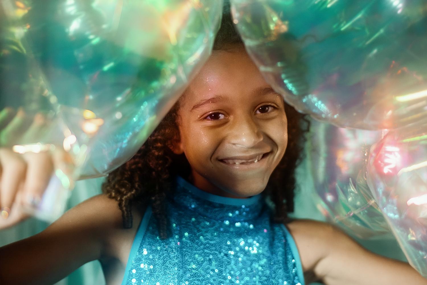 Smiling young girl in blue sequin top peeking through shiny iridescent plastic wrap with colorful light reflections.