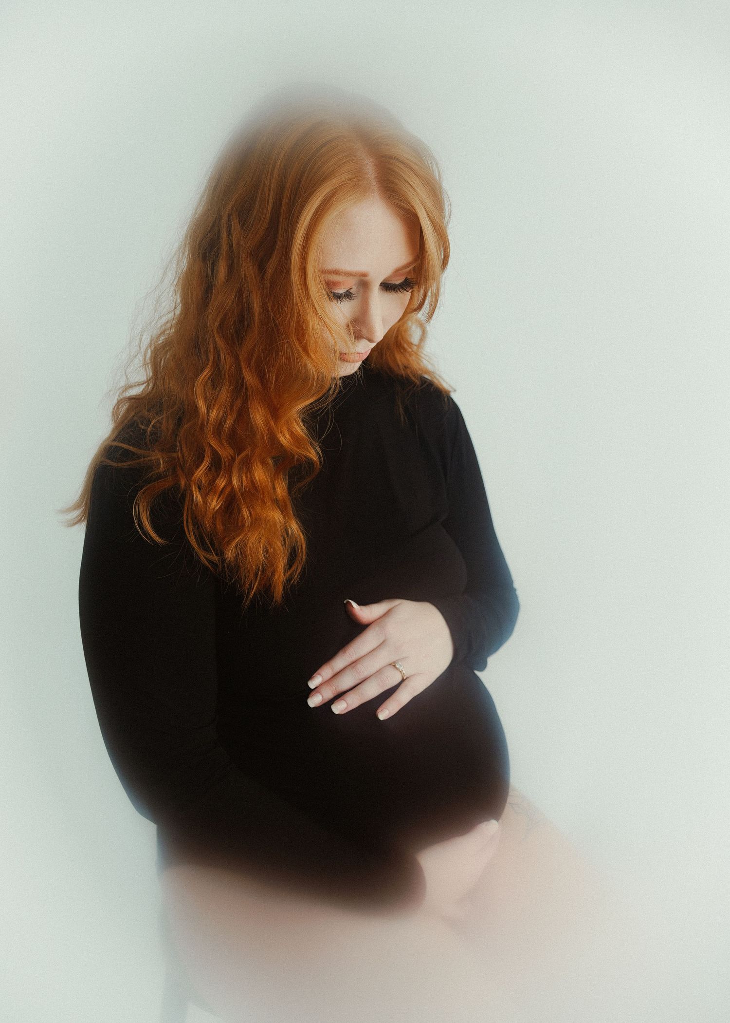 Artistic maternity photo of a woman in black dress with flowing red hair against light background.