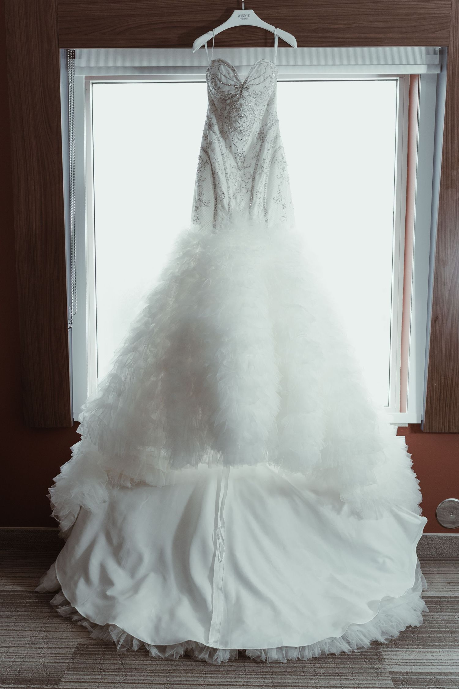 An elegant white wedding dress with a full tulle skirt hangs in front of a window.