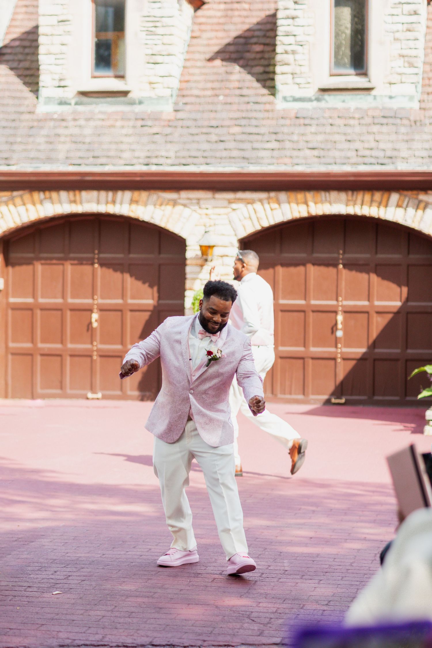 Wedding guest in light pink suit waves to guests while walking down outdoor aisle.