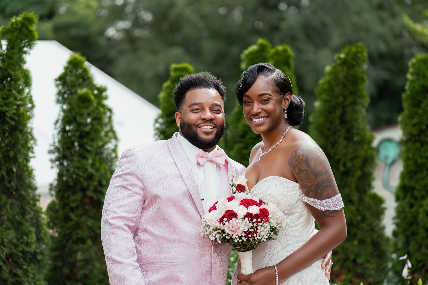 Newlyweds pose with a red and white bouquet in front of lush greenery at their outdoor celebration.