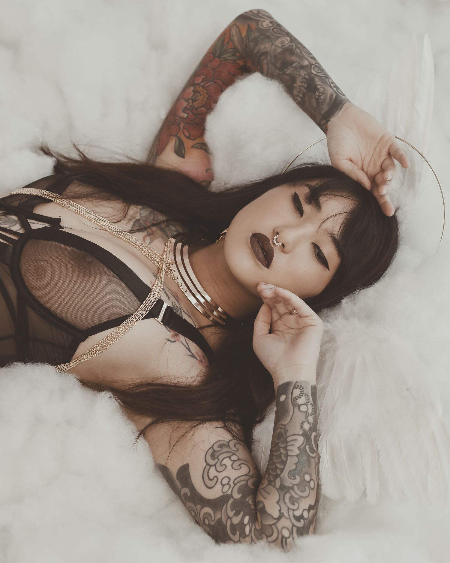 Artistic boudoir photo with tattooed model posed among white clouds wearing dark lingerie.
