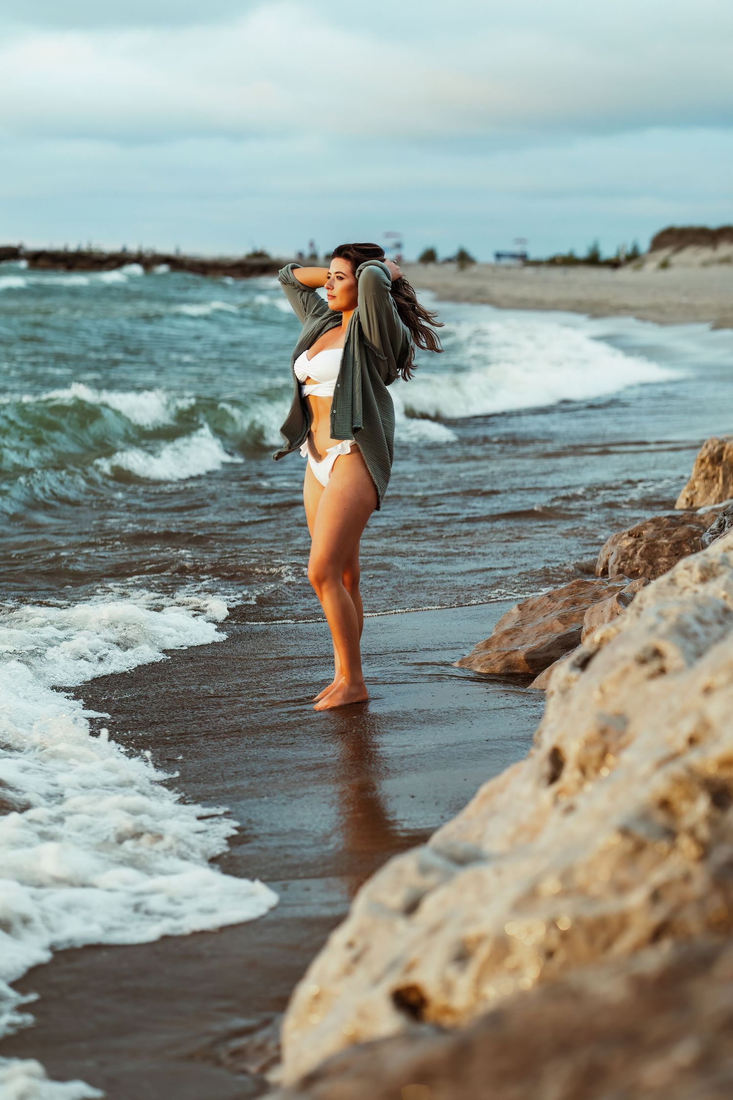 Woman in swimwear and cardigan standing on rocky beach with waves crashing.