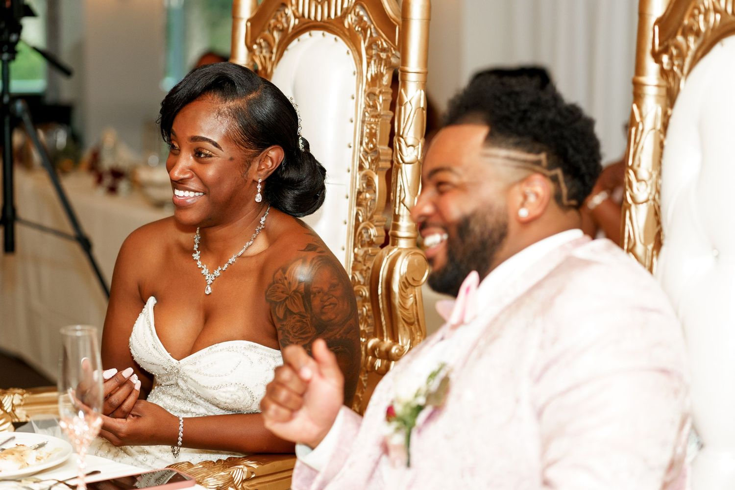Newlyweds seated at ornate gold thrones share a happy moment during their reception.