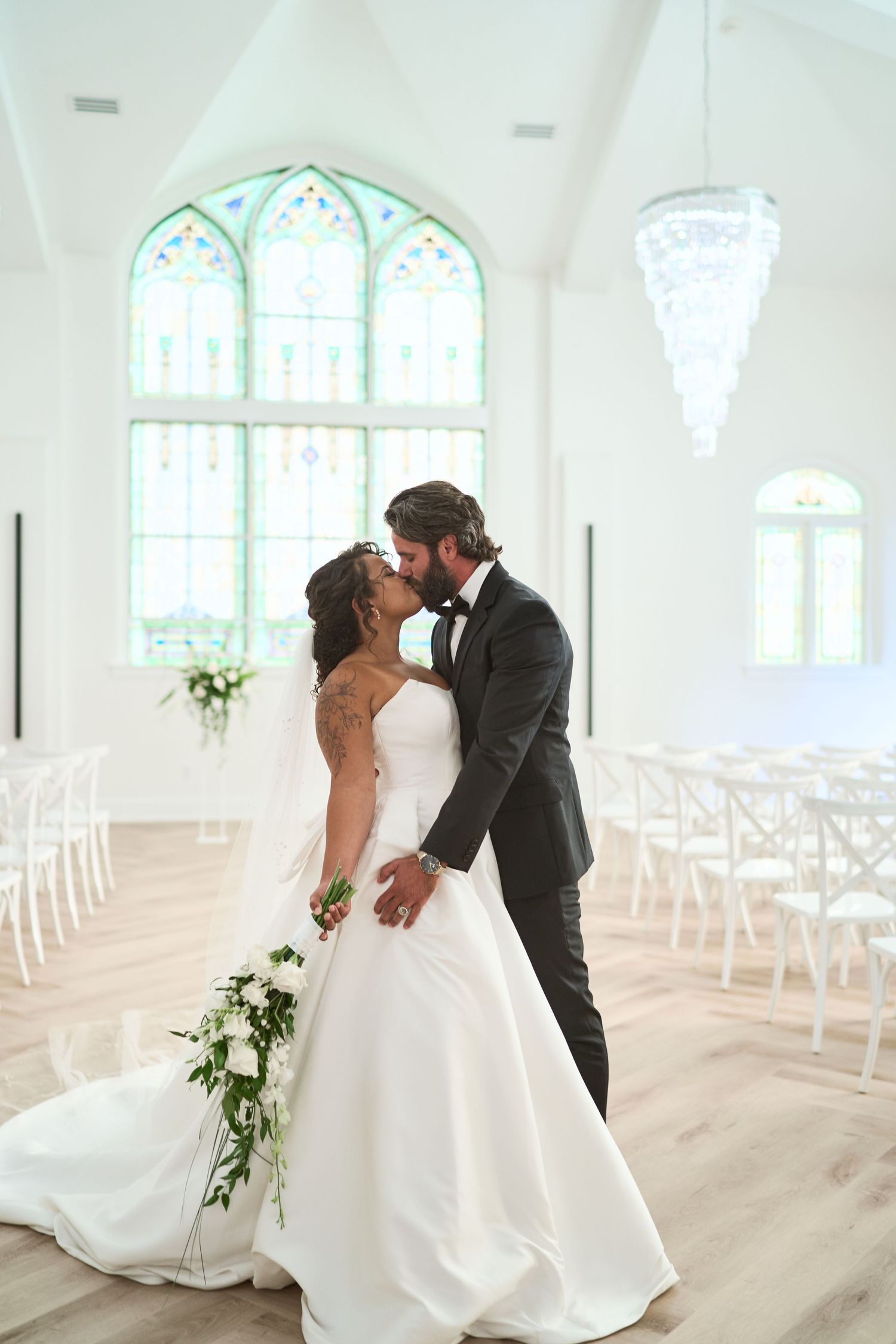 A romantic wedding moment in a bright chapel with stained glass windows and hanging crystal chandeliers.