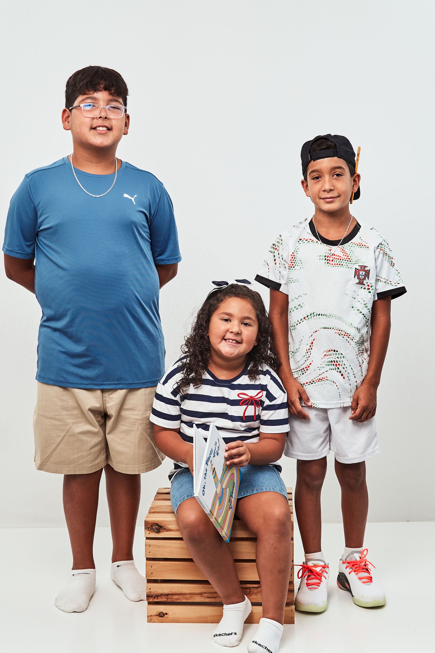 Three children in casual outfits pose together near wooden blocks against a white studio background.