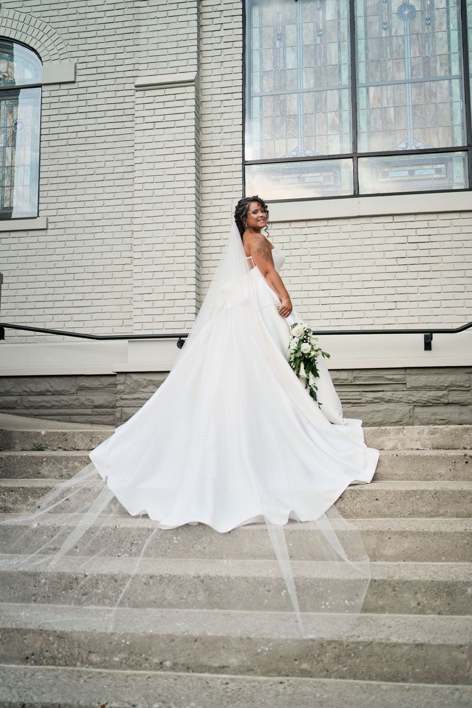 A bride in a flowing white wedding gown poses gracefully on stone steps in front of a white brick church.