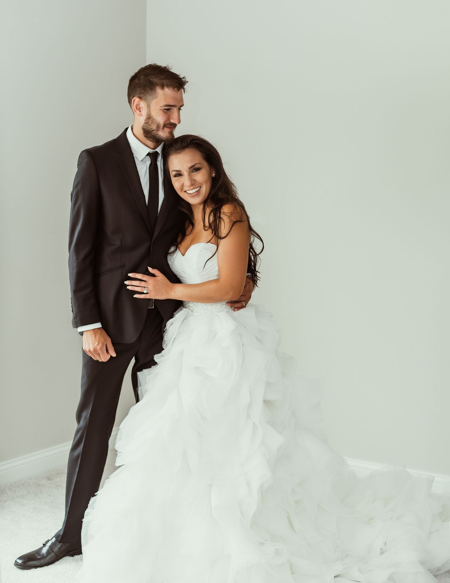 A bride in a flowing white dress embraces her groom in a dark suit during their wedding photoshoot.