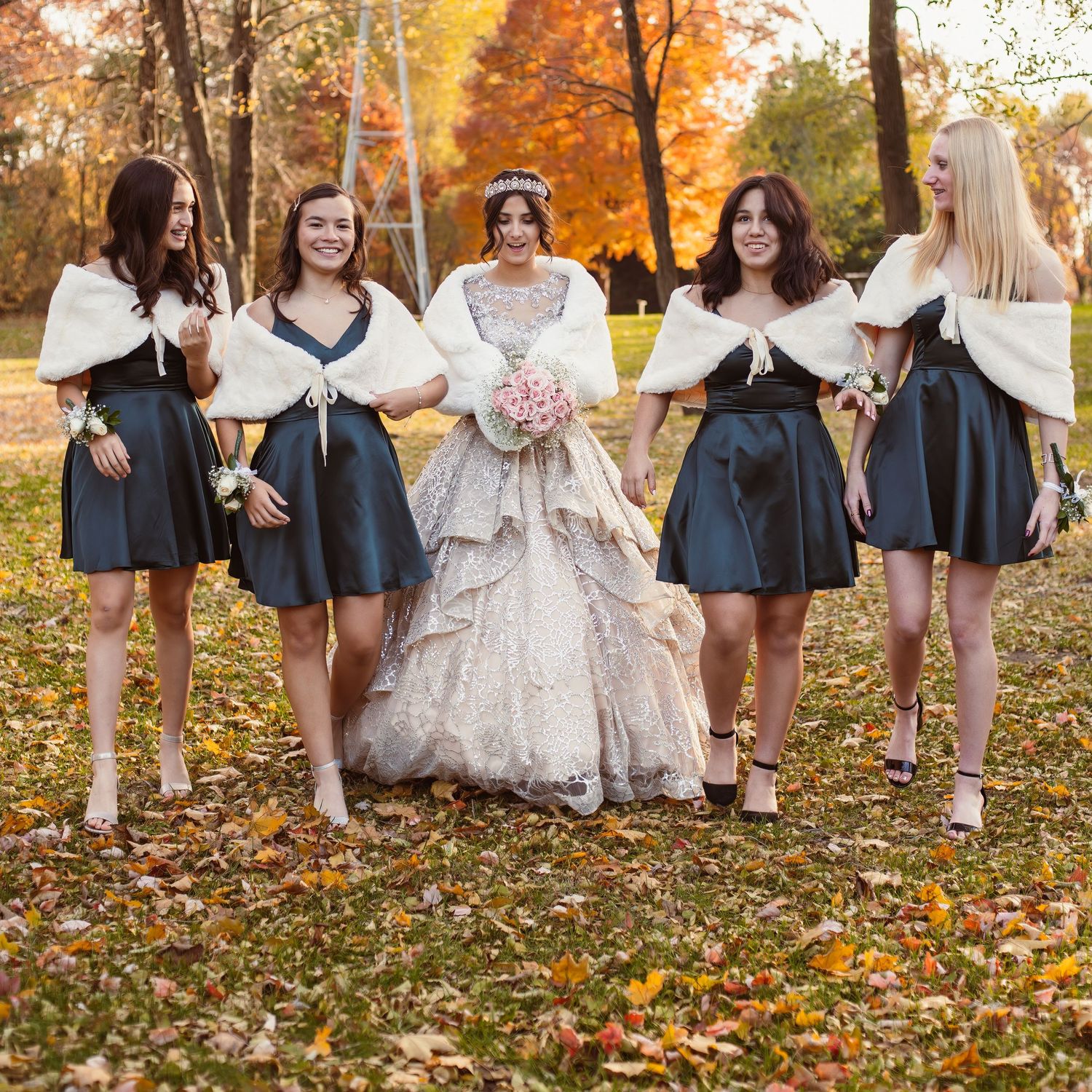 Bridal party in navy dresses and white shawls poses with bride in autumn setting.