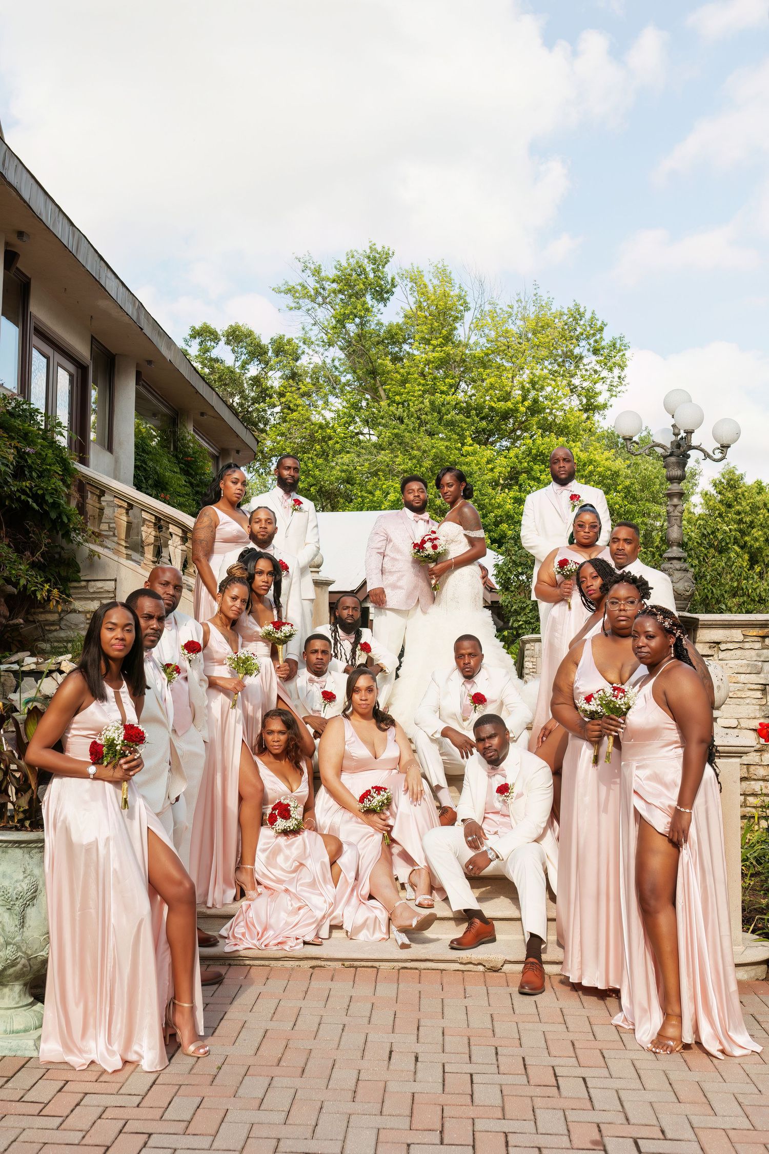 Large wedding party in pink dresses and white suits poses on elegant stone staircase.