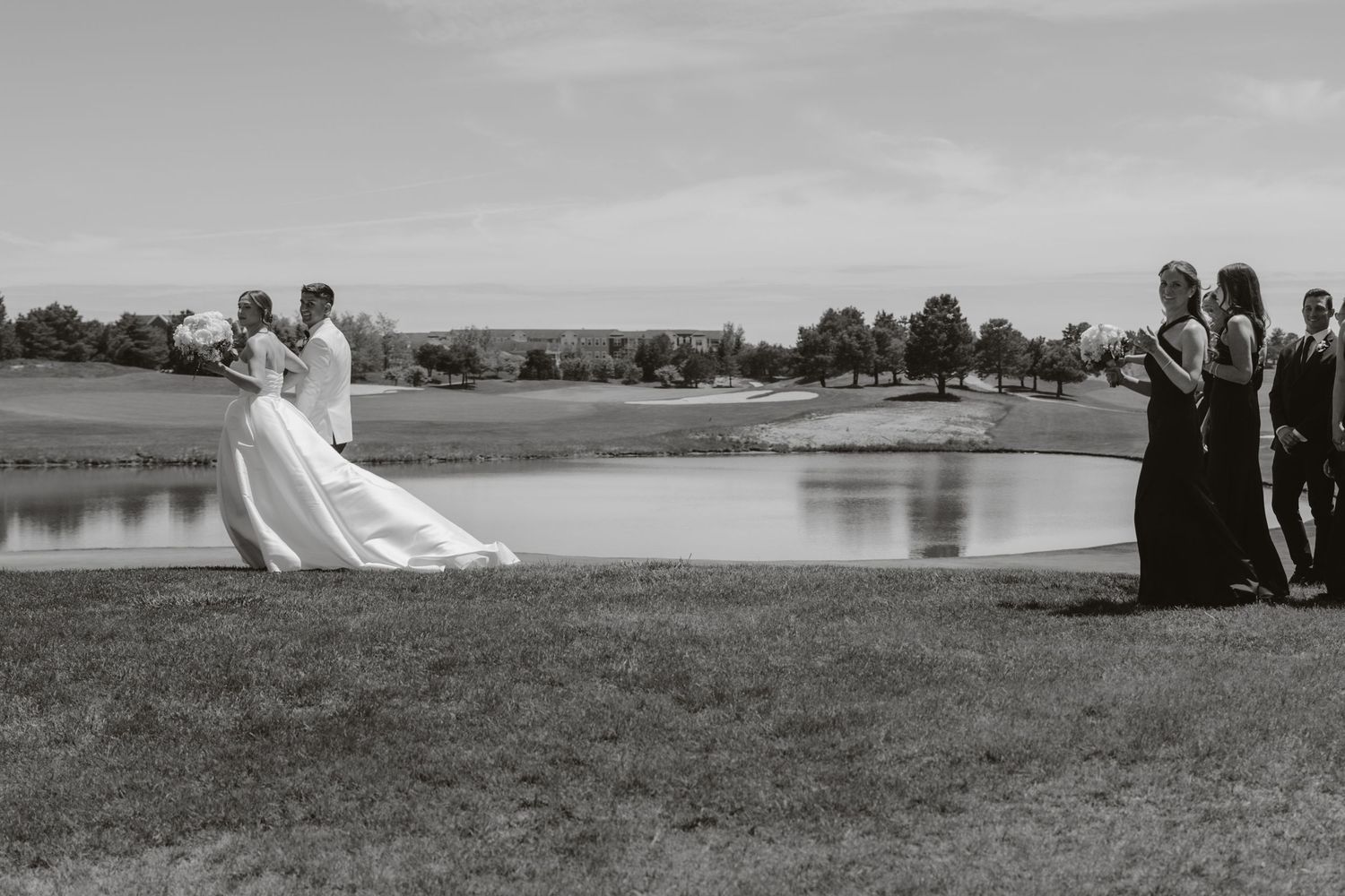 Black and white wedding party photo taken on a grassy hill overlooking a serene lake with trees in the background.