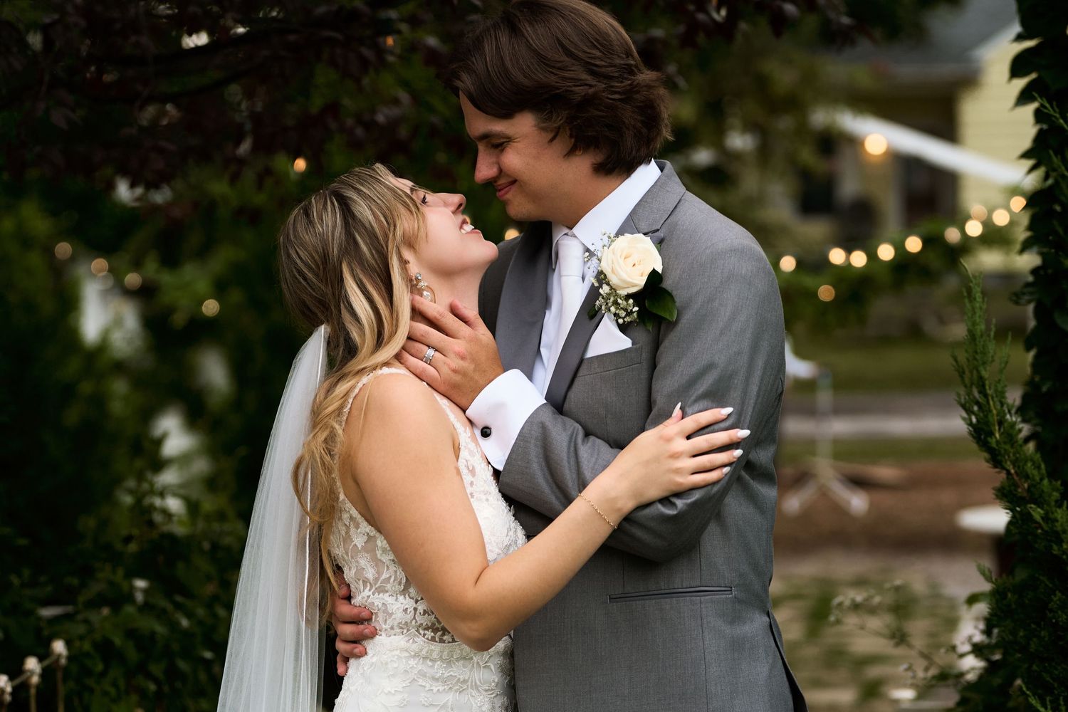 A couple shares a romantic kiss during their outdoor wedding celebration with string lights in the background.