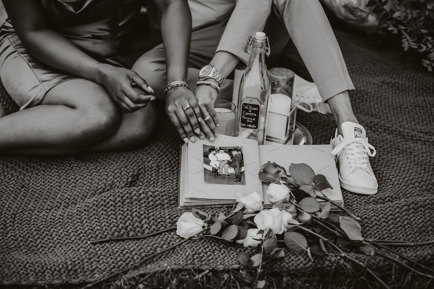Memory-filled picnic scene with photo album, drinks and flowers on woven blanket.