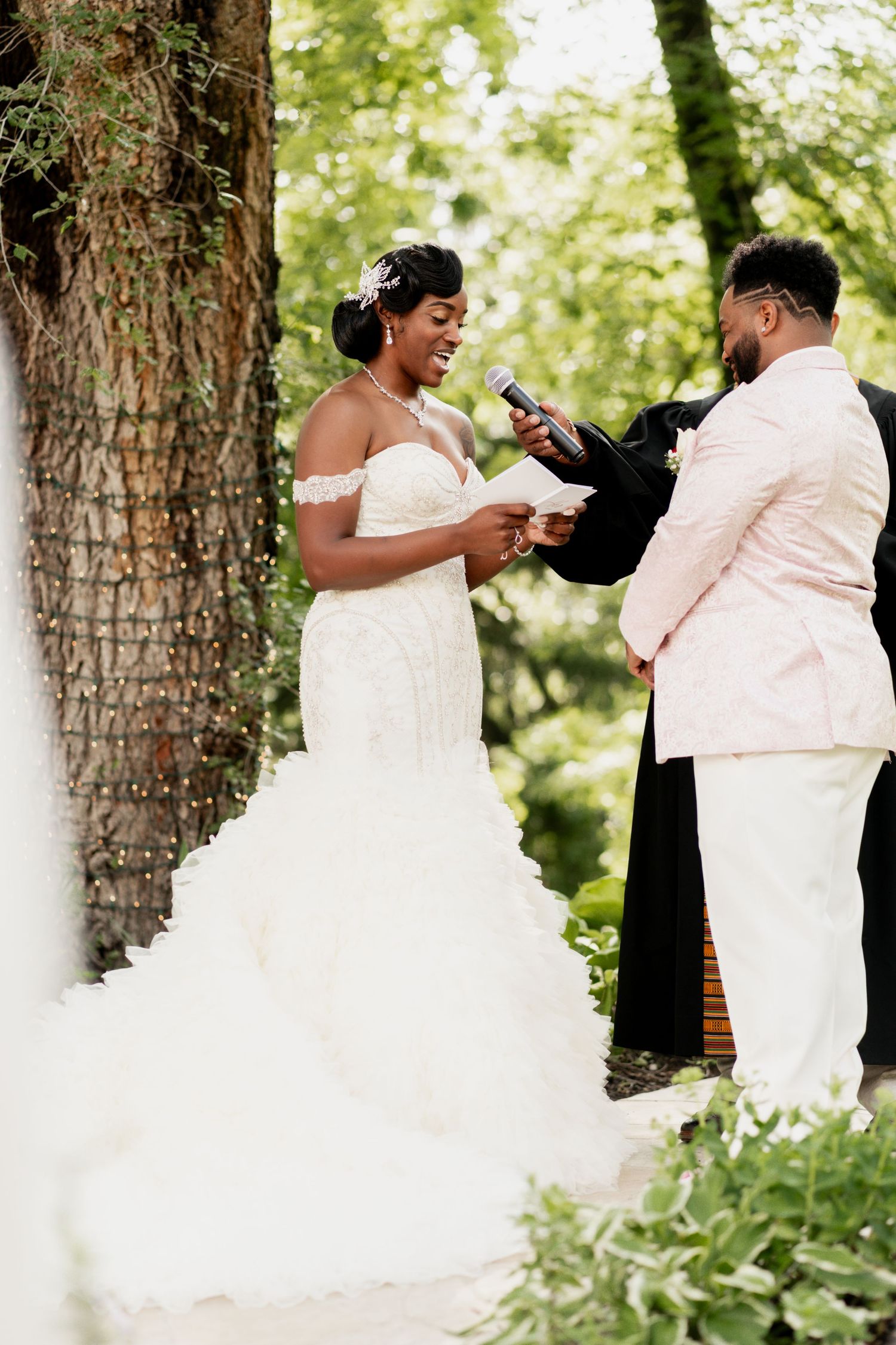 A couple exchanges rings during their outdoor wedding ceremony near a large tree.