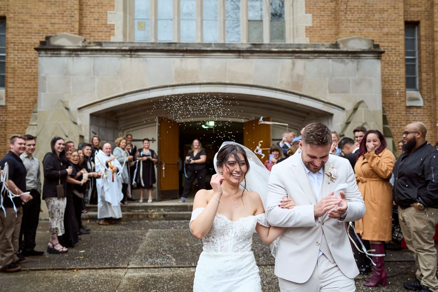 A joyful couple exits a church with brick facade while wedding guests applaud and celebrate their union.