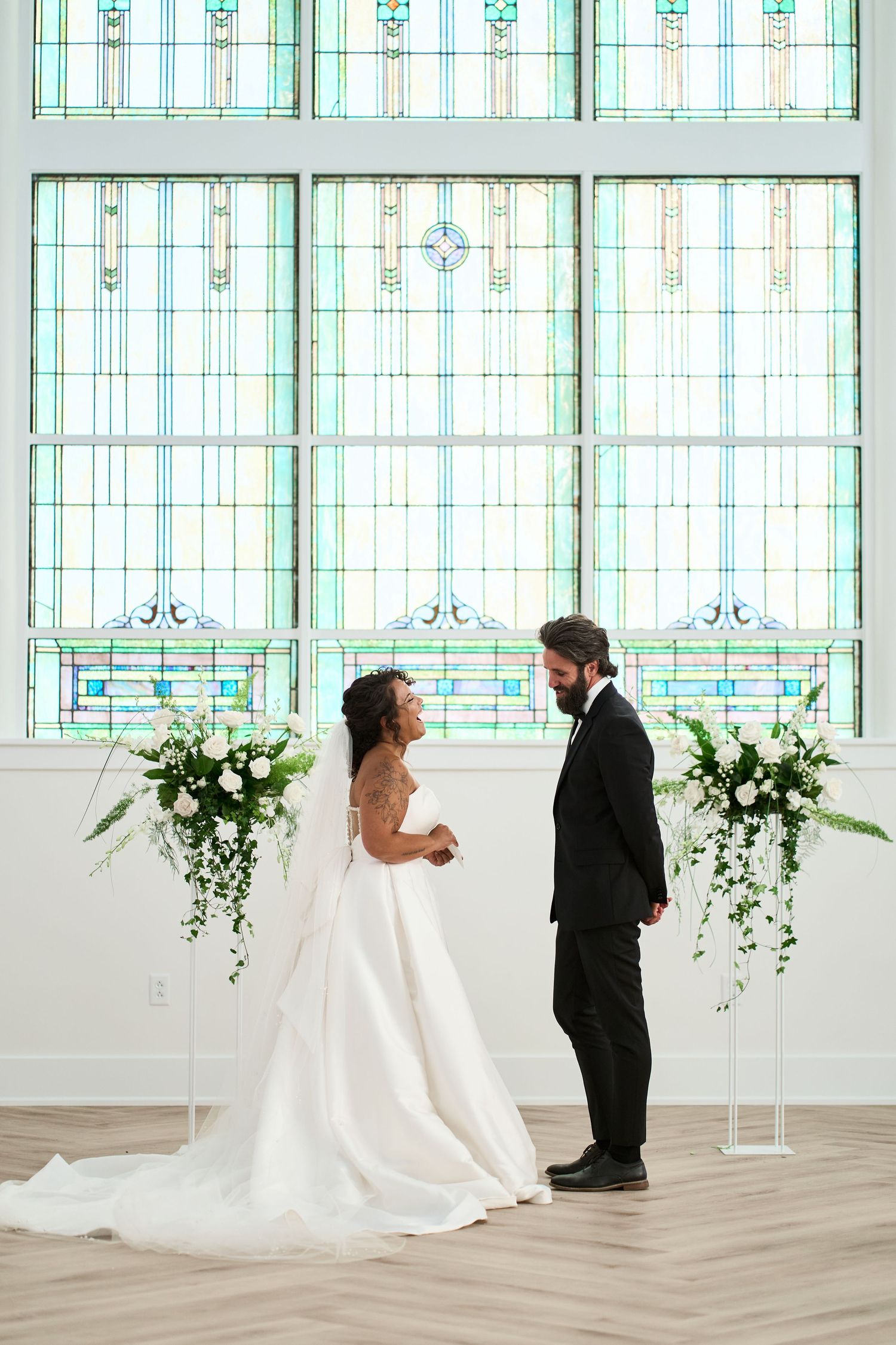 A couple exchanges vows in front of magnificent stained glass windows with white floral arrangements.