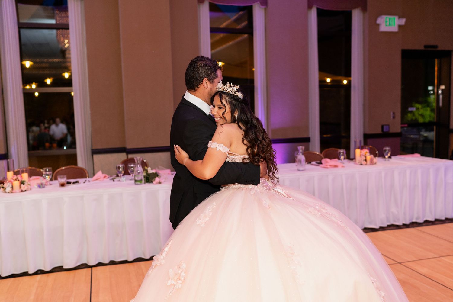 A couple shares their first dance at a wedding reception with a white-decorated banquet hall in the background.