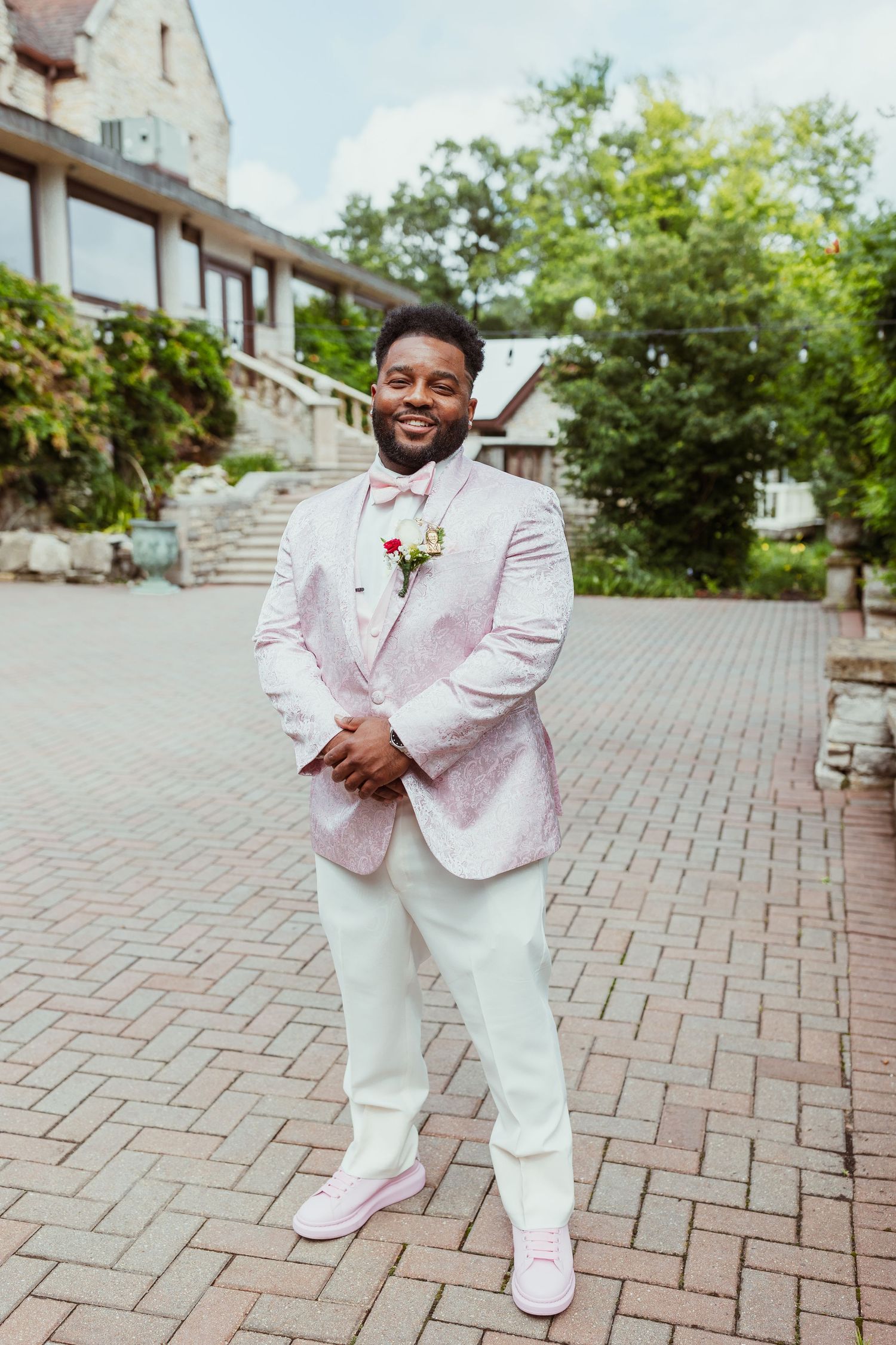 Groom in a stylish pink and white suit with matching shoes poses on brick pavers outdoors.