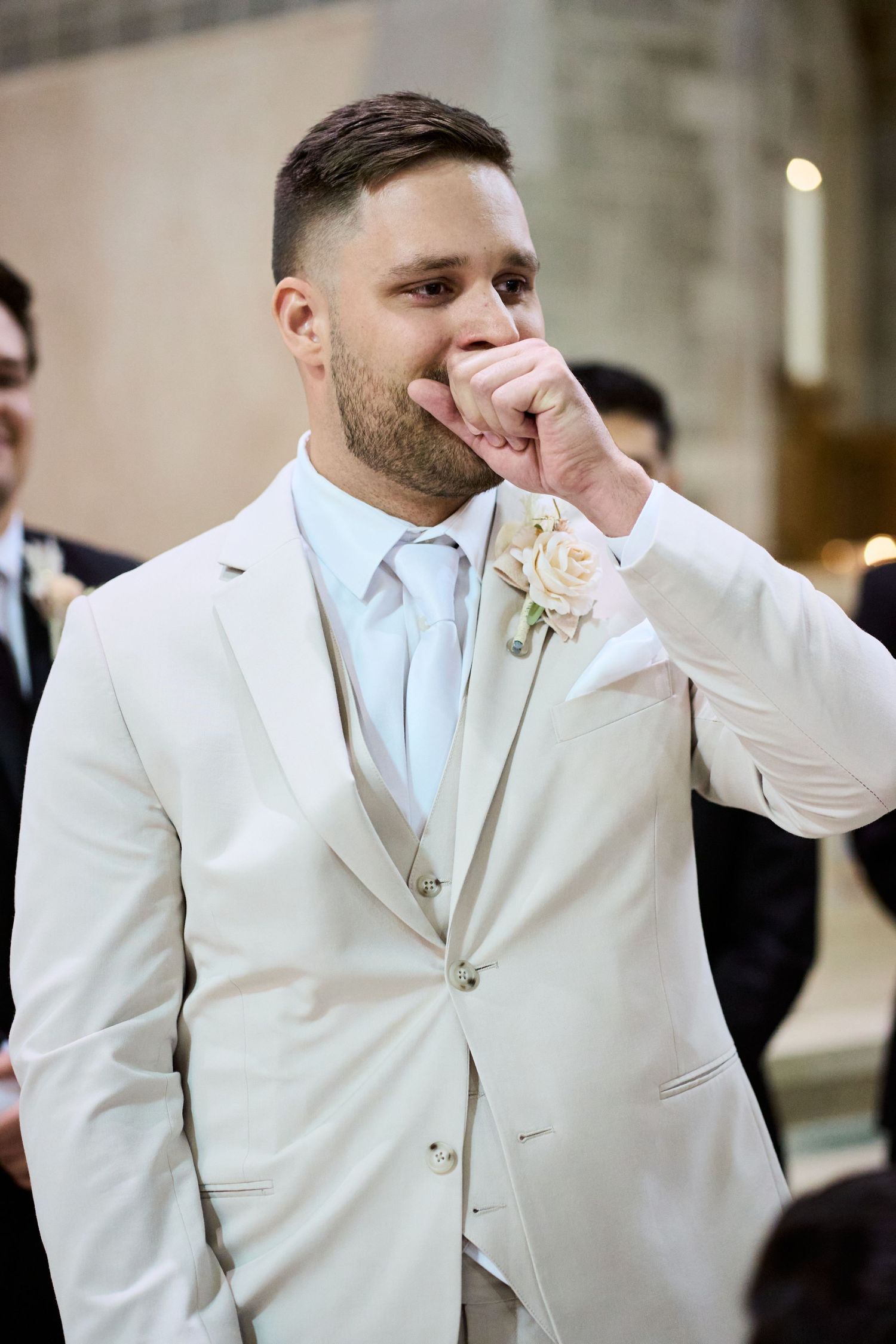Emotional groom in white suit and tie wipes away tears during wedding ceremony.