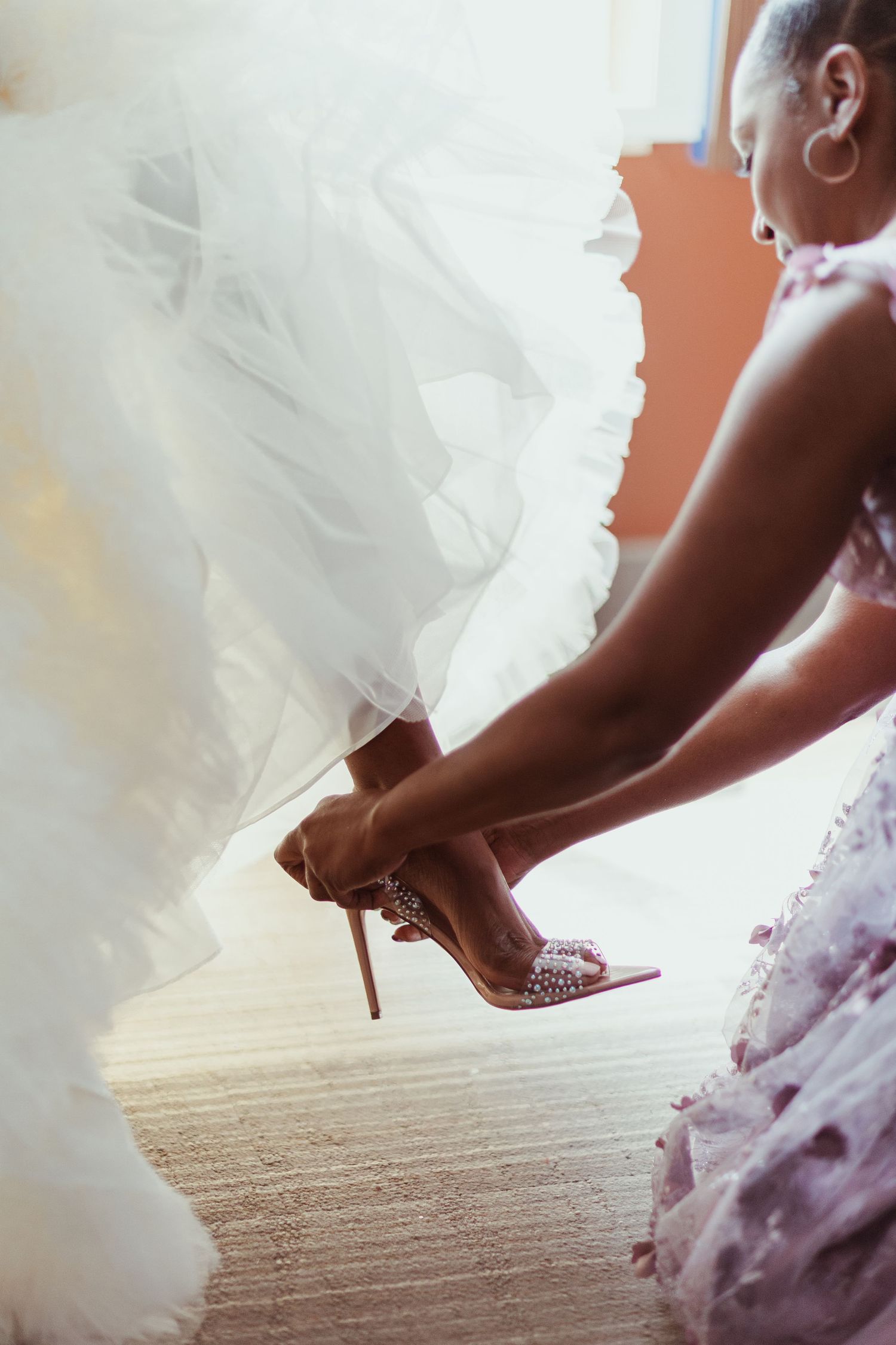 Someone helps adjust sparkly bridal shoes beneath layers of white tulle wedding dress.