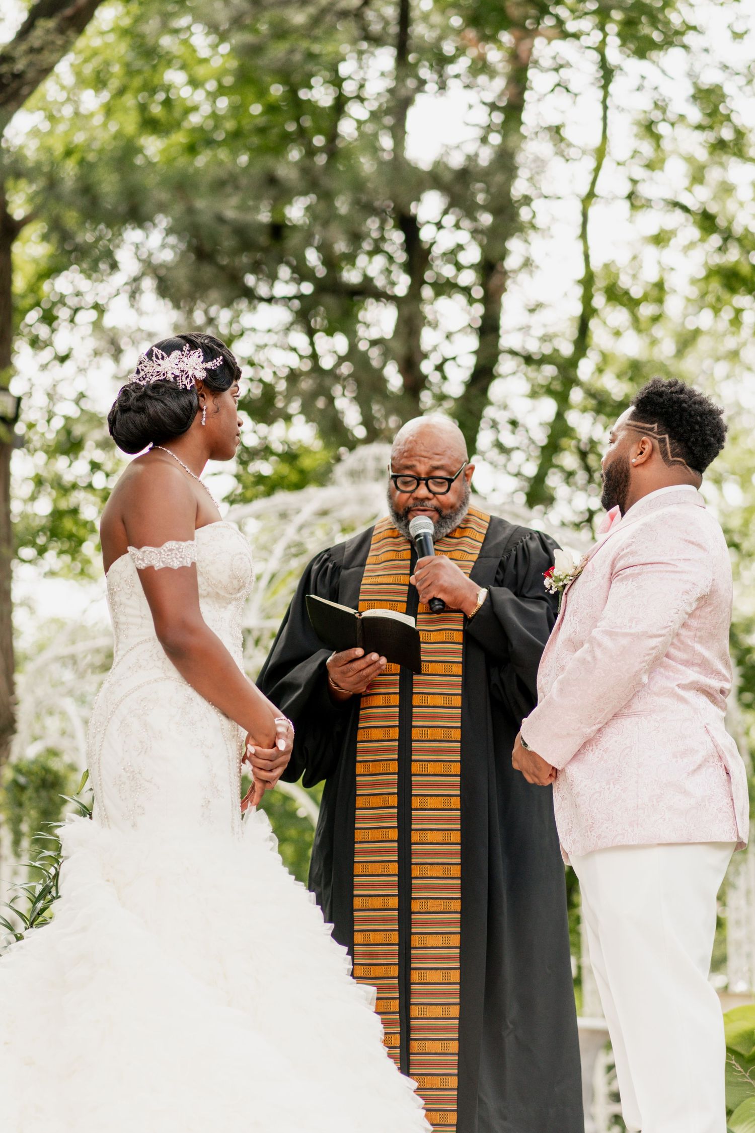 A wedding ceremony takes place outdoors with participants standing before an officiant wearing traditional robes.
