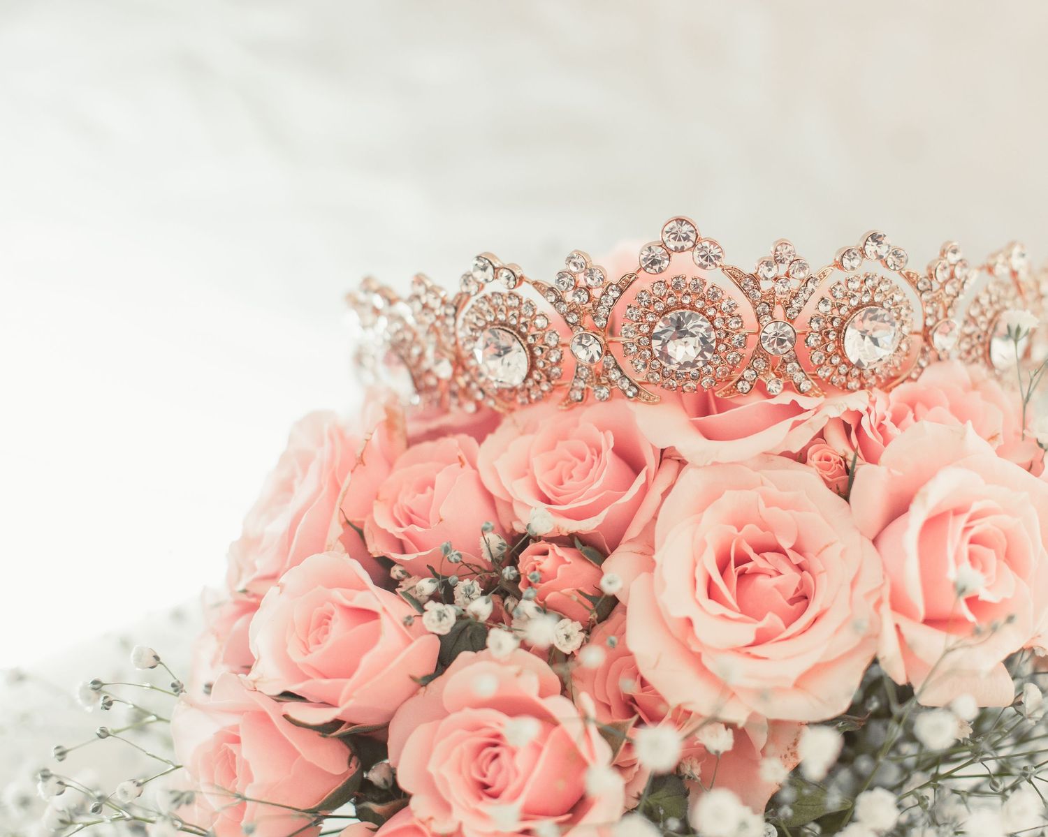 Rose gold tiara with crystals resting on pink roses and baby's breath flowers.