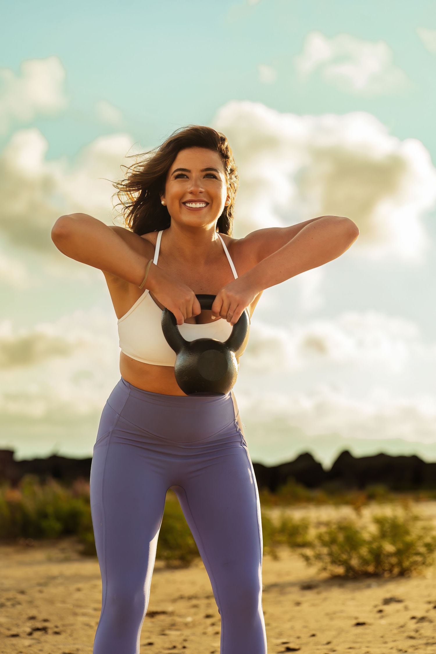 Woman performs kettlebell exercise on beach with bright sky background.