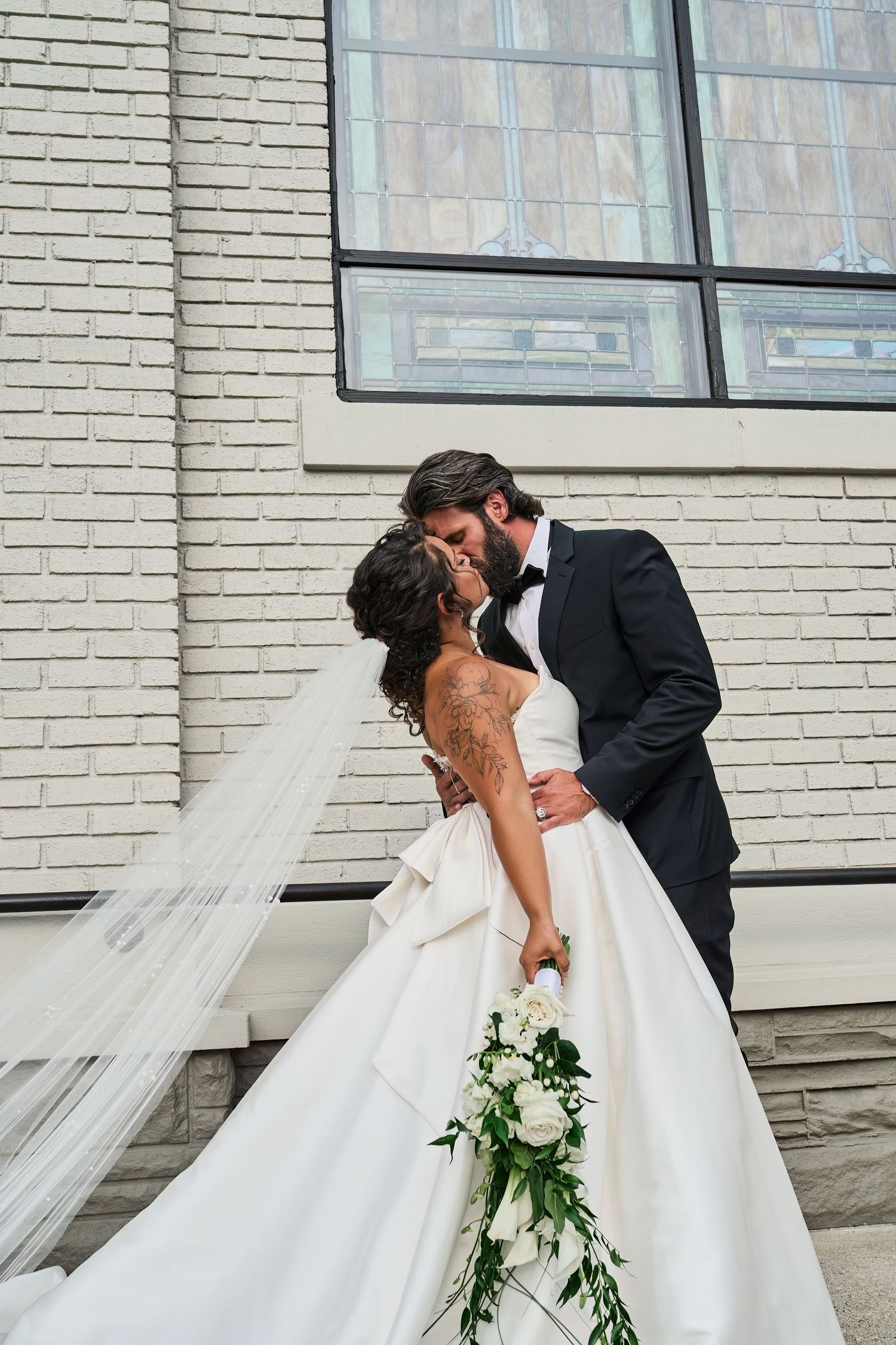 Elegant newlyweds share a romantic kiss against a brick wall as the bride's veil flows in the breeze.