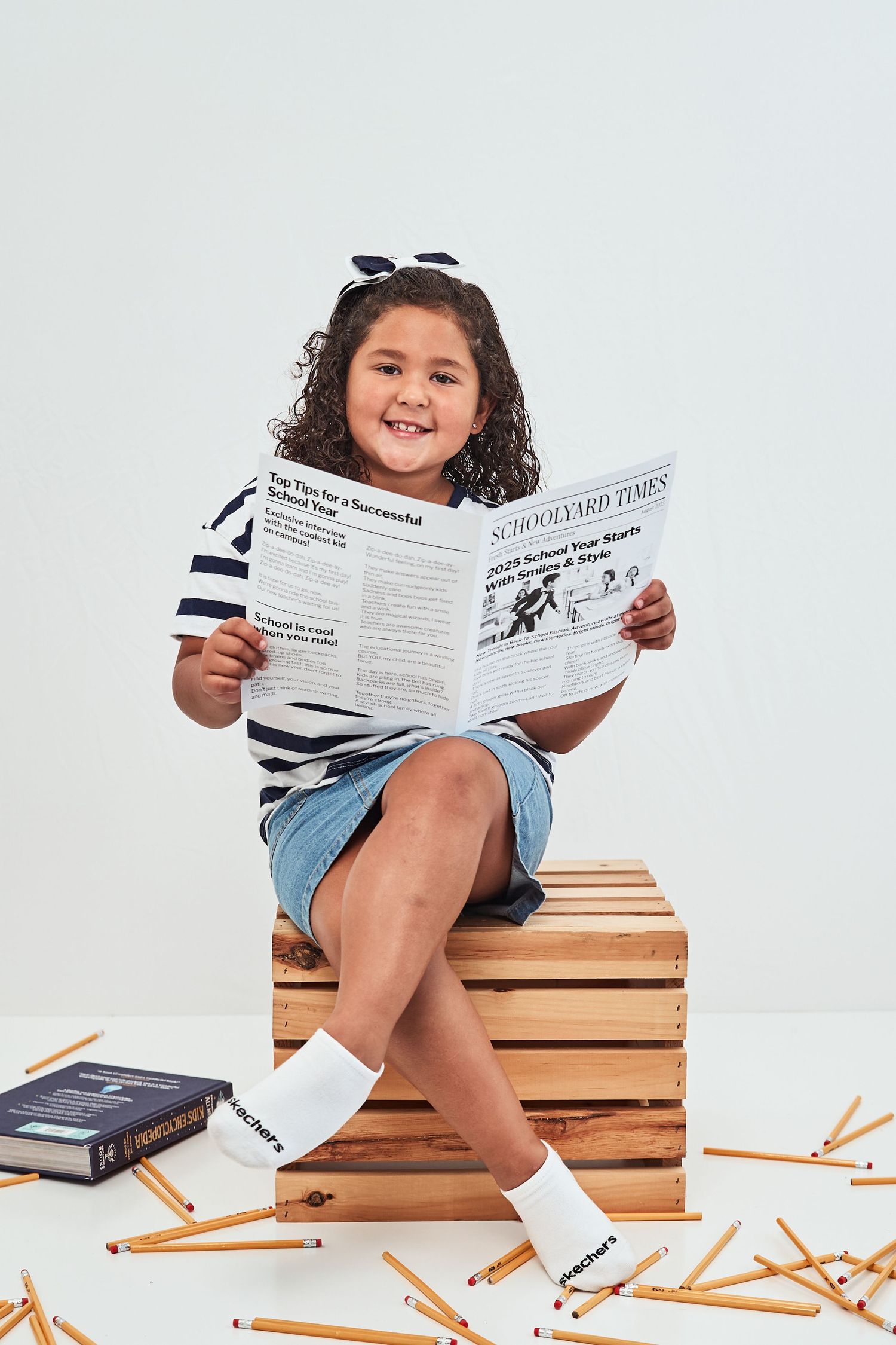 A child sits on a wooden crate reading a document with pencils scattered on the floor.